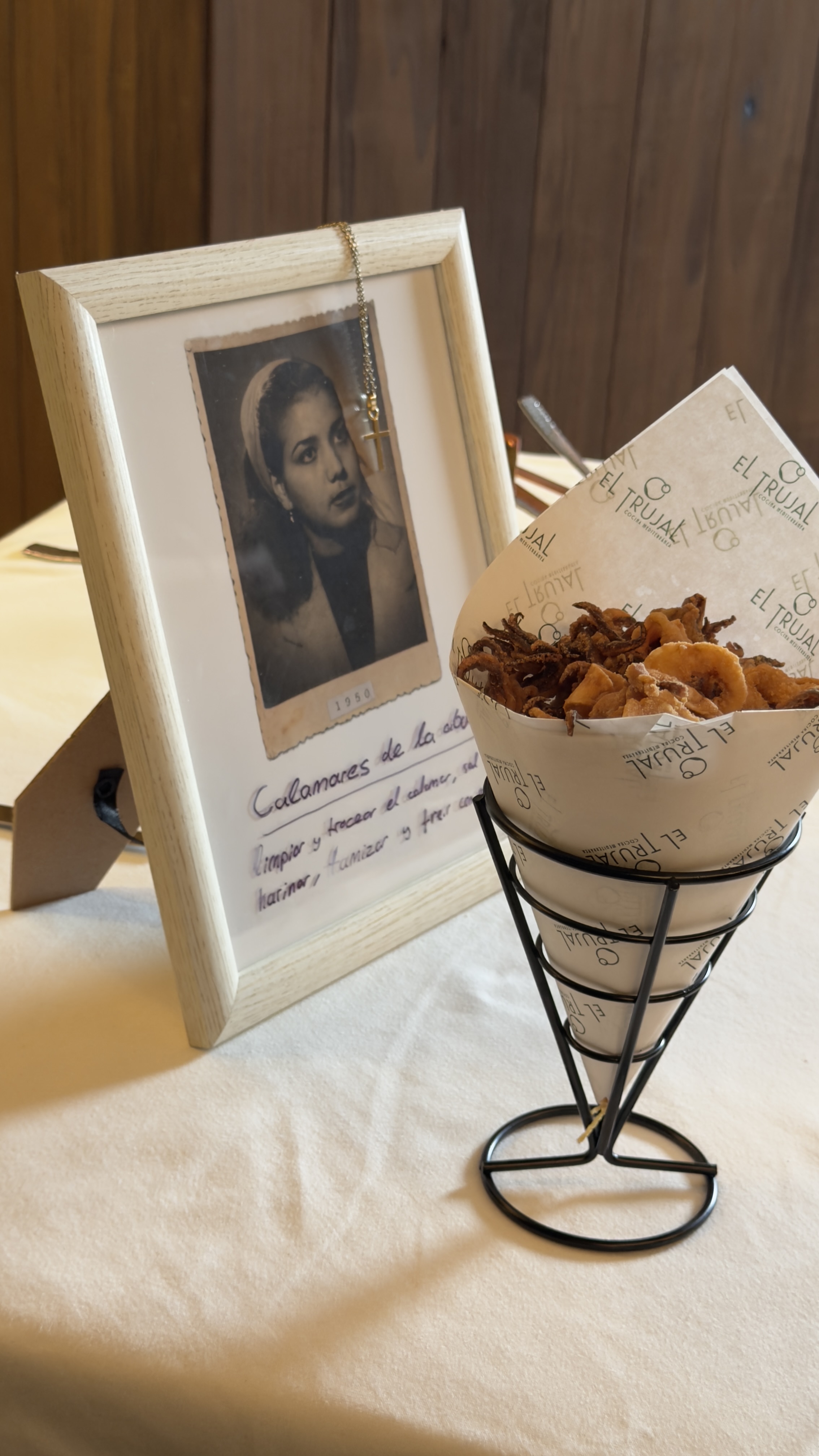 La mesa en El Trujal: retrato de la abuela enmarcado con la receta manuscrita de calamares, al lado un cucurucho con la tapa servida por el restaurante